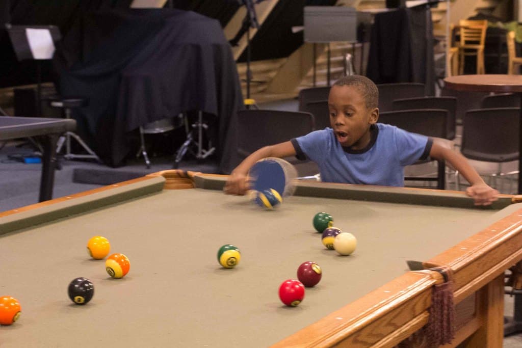 Ein Junge hält einen Tischtennisschläger in der Nähe eines Snookertisches mit Billardkugeln, mit Stühlen und anderen Gegenständen im Hintergrund.