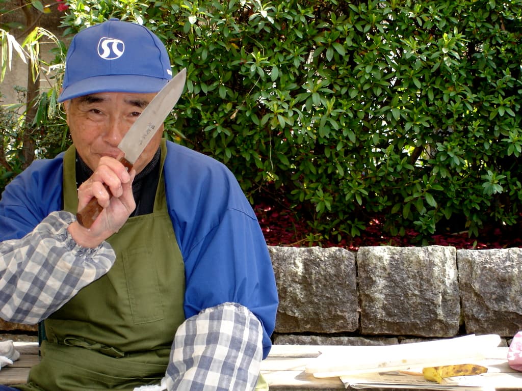Ein älterer Mann in einer Schürze hält ein Messer und steht vor Papieren mit Felsen und Bäumen im Hintergrund.