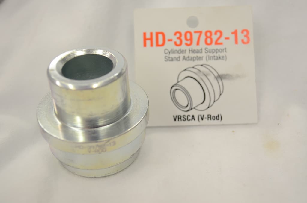 Ein Metallobjekt mit einer Beschriftung, die 'HD 397 82 13 Zylinderkopfträgerstandadapter Einlass' lautet.