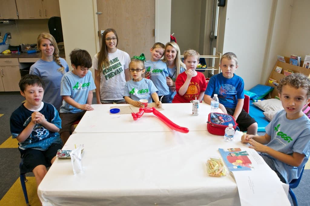 Eine Gruppe von Kindern und drei Frauen, einige lachend, mit Schränken, einer Wand, einer Tür und Kissen im Hintergrund.