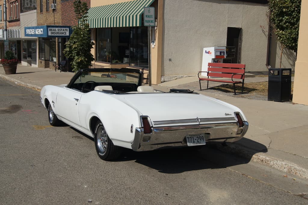 Ein weißes Auto mit der Nummer 771-299 steht auf einer Straße, rechts daneben eine Bank, im Hintergrund ein Gebäude und ein Geschäft namens "Immobilienversicherung".