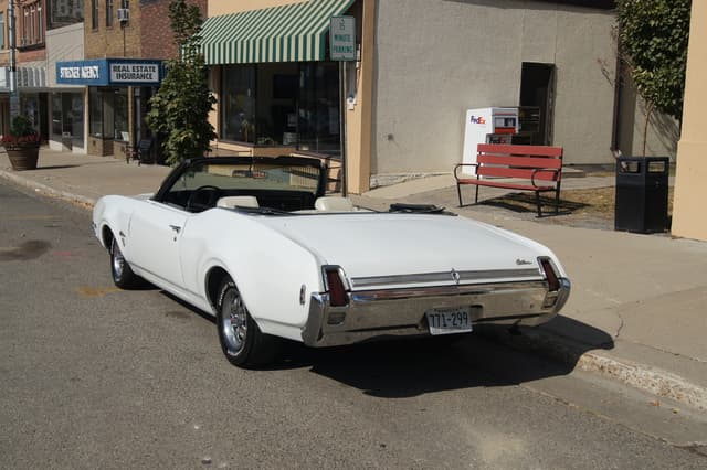 Ein weißes Auto mit der Nummer 771-299 steht auf einer Straße, rechts daneben eine Bank, im Hintergrund ein Gebäude und ein Geschäft namens "Immobilienversicherung".