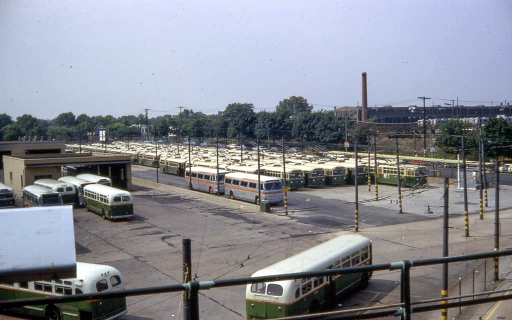 Eine Reihe von parkenden Bussen auf einer Straße mit Polen, Drähten, Gebäuden, Bäumen, einem Zaun, einem Rauchschlot und einem Himmel im Hintergrund.