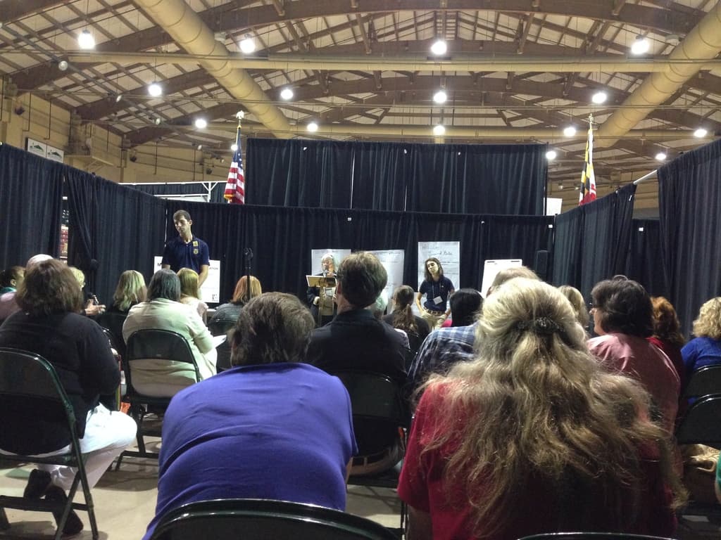 Eine Gruppe von Menschen, die auf Stühlen sitzen, mit einer Person, die an einem Rednerpult steht, vor einem Hintergrund aus Bannern, blauen Vorhängen, Flaggen und Deckenleuchten.