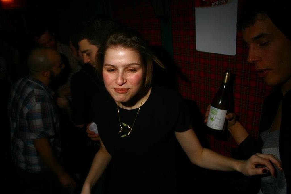 Eine Gruppe von Menschen in einem Raum, mit einer Frau in einem schwarzen Kleid vorn, einem Mann in einer schwarzen Jacke mit einer Flasche rechts und einem roten Tuch im Hintergrund.