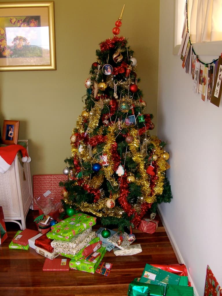 Ein geschmückter Weihnachtsbaum rechts mit Geschenken darunter, ein Foto Rahmen an der linken Wand und ein Vorhang am rechten Fenster.