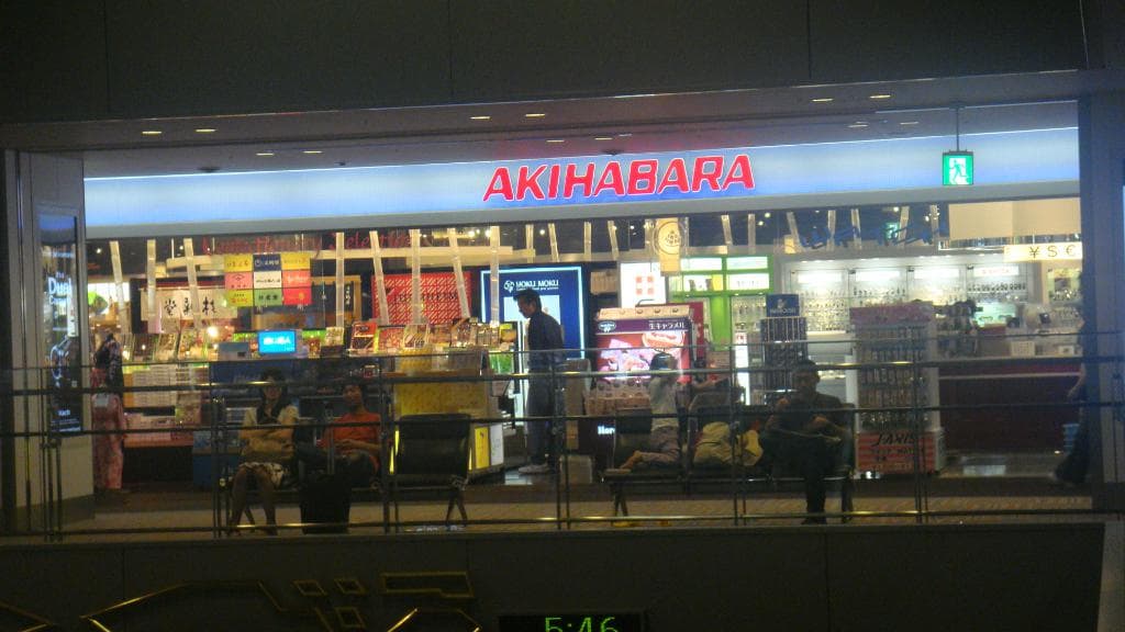 Ein Geschäft mit zwei Bänken davor, Menschen darauf sitzend, eine Wand mit Text darüber, Tabletts mit Gegenständen darin und eine eingezäunte Wand unten.