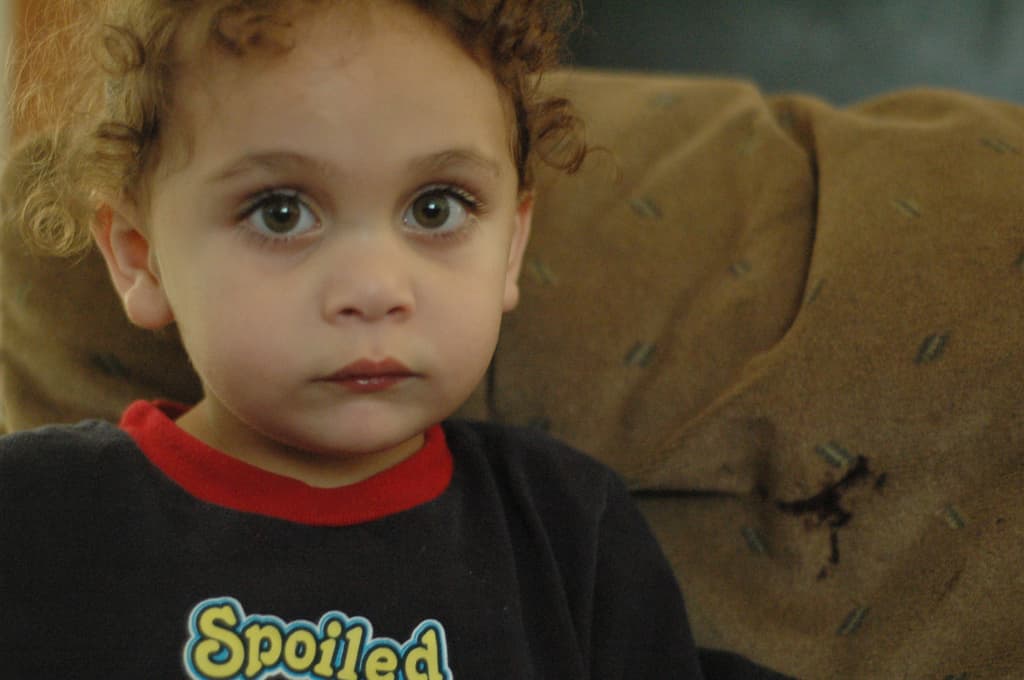 Ein Kind mit lockigen Haaren, das ein schwarzes Hemd mit der gelben Aufschrift 'Verwöhnt' trägt, vor einem braunen Stoffhintergrund.