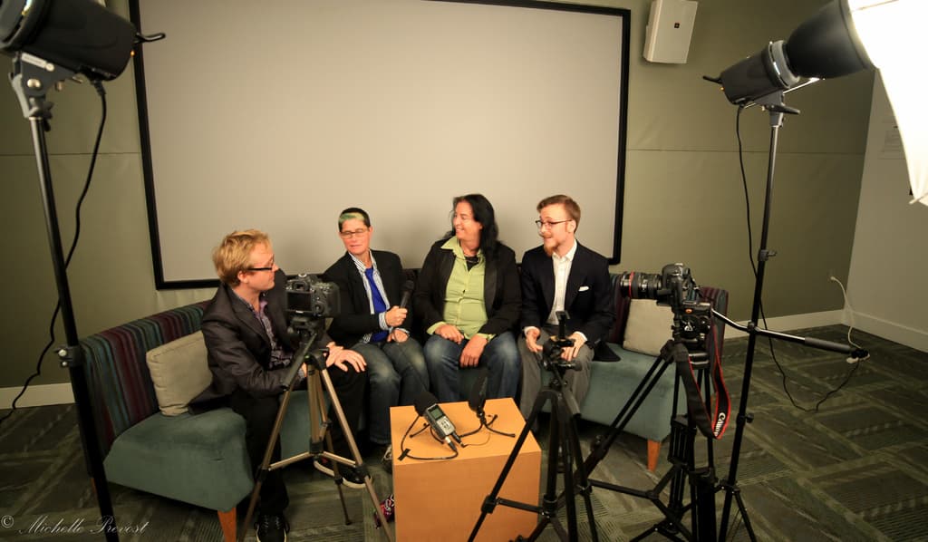 Eine Gruppe von drei Personen sitzt auf einer Couch und gibt ein Interview. Links sitzt eine Person in einem schwarzen Mantel und Hose, in der Mitte eine Person in einem Mantel und blauer Krawatte mit einem Mikrofon und rechts eine Person, die strahlt. Auf dem Tisch in der Mitte steht ein Mikrofonrekorder, Kameras sind auf der rechten und linken Seite und große Scheinwerfer sind vorhanden, im Hintergrund ein Projektionsschirm.