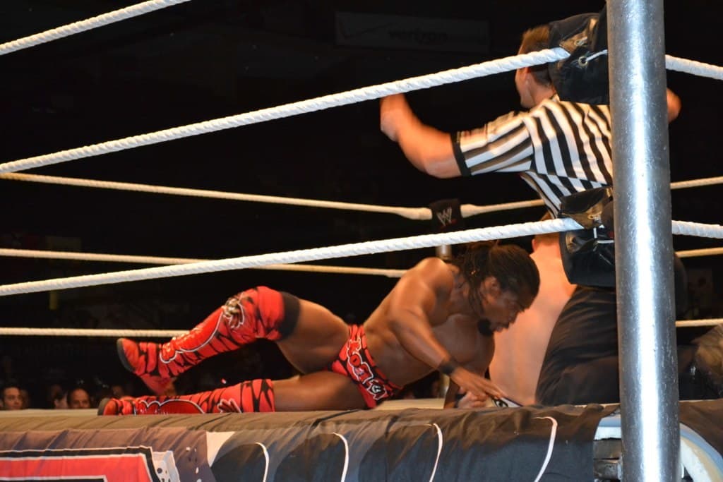 Zwei Männer in einem Boxring, einer in einem roten Hemd und Schuhen, der andere in Shorts, kämpfen, mit einem Schiedsrichter rechts und einer Zuschauermenge im Hintergrund unter einem dunklen Himmel.