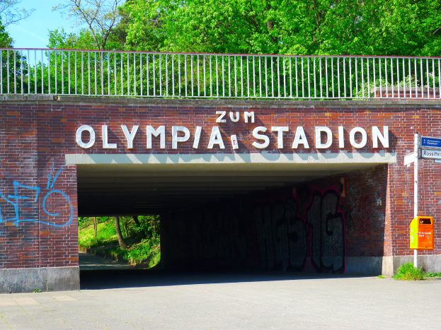 Der Eingang zum Olympiastadion in Berlin, Deutschland, mit einer Br%C3%BCcke mit Text, einem Metallzaun, einer Tafel, einer Kiste, Pflanzen, Gras, einer Baumgruppe und einem bewölkten Himmel.