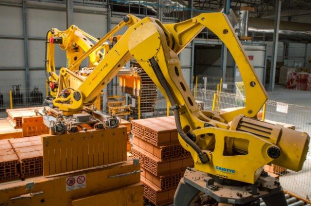 Ein großes Lager mit zahlreichen Kisten, Kränen, einem Zaun, Pfählen und anderem industriellen Equipment, mit einer Wand im Hintergrund, die eine größere industrielle Einrichtung andeutet.