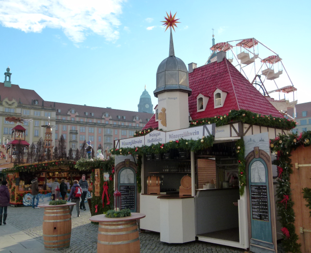 Ein geschäftiger Weihnachtsmarkt in Nürnberg, Deutschland, mit Menschen um Stände, die mit Lichtern und Schmuck verziert sind, Gebäuden im Hintergrund, einem Riesenrad und einer Tafel mit Text auf der rechten Seite.