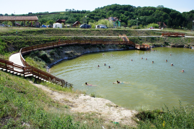 Gruppe von Menschen, die in einem Gewässer schwimmen, mit üppiger Vegetation, einer Brücke mit Treppen, Hütten, Fahrzeugen, Pfählen und einem klaren blauen Himmel im Hintergrund.