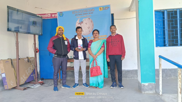 Eine Gruppe von vier Personen steht vor einem Gebäude, in der Mitte eine Frau mit einer Tasche, zwei Männer in Jacken daneben und links ein Fernseher auf einem Pfahl mit verstreuten Gegenständen drumherum; im Hintergrund Schilder mit Text, eine blaue Tür, eine Wand, ein Fenster und ein Geländer, feiernd ihren Sieg in der ersten Runde der Saison 2018-19 des Chhattisgarh Board of Secondary Education.