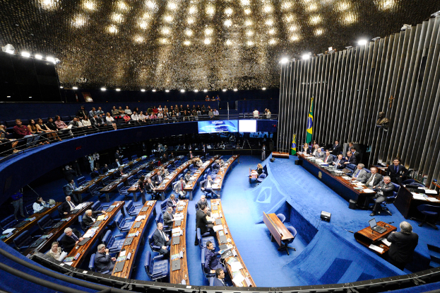 Eine Konferenzbühne mit sitzenden Personen an einem Tisch mit Papieren und Gegenständen, flankiert von Fahnen und einer Skulptur, mit Gästen an Tischen und hinter einer Absperrung, beleuchtet von Deckenleuchten und zwei Bildschirmen.