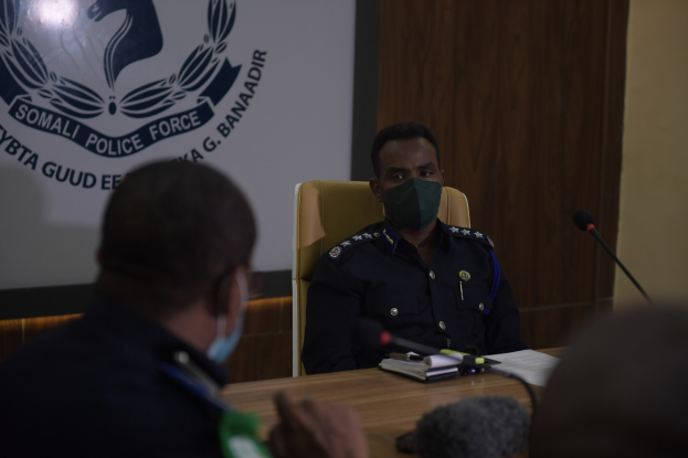 Zwei Polizeibeamte mit Masken sitzen an einem Tisch mit Mikrofonen, Büchern und anderen Gegenständen und schauen auf einen Wandbildschirm mit Text und einem Emblem.