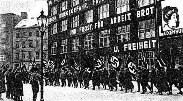 Schwarzes und weißes Foto einer Gruppe, die mit Fahnen und Transparenten die Straße entlangmarschiert, mit einem Laternenpfahl links und einem Gebäude mit der Aufschrift "Deutsche Nationalsozialistische Arbeiterpartei in Berlin" rechts.