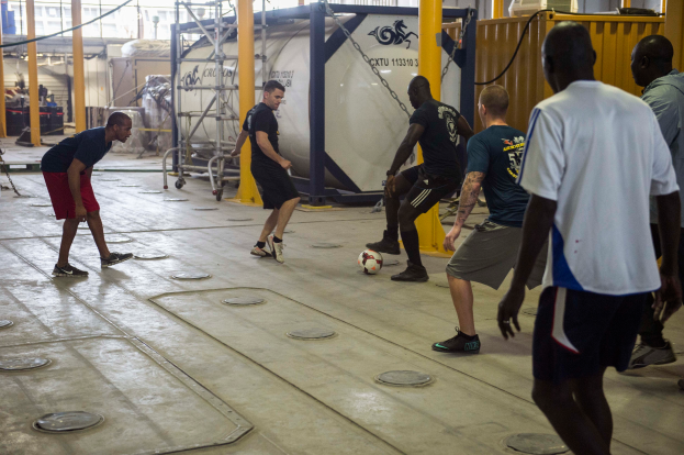 Eine Gruppe von Männern, die drinnen Fußball spielen, mit einem Ball auf dem Boden, Metallstangen, einem Behälter, einem Metallzaun, Stangen mit Ketten, einer Wand und einer Decke mit Lampen.