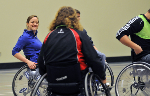 Eine Gruppe von Menschen in Rollstühlen auf dem Boden sitzend, mit einer Frau in einer blauen Jacke und einer Person in einem grünen Hemd, vor einer Wand im Hintergrund.