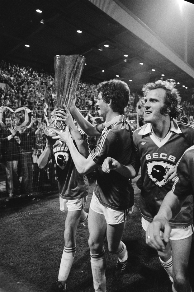 Schwarzes Bild von Fußballspielern, die mit einem Pokal auf einem Feld feiern, umgeben von einer Menge hinter einem Zaun unter Stadionbeleuchtung.