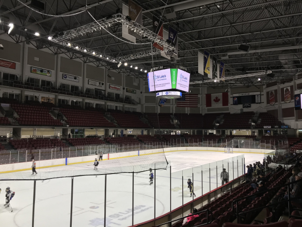Eishockeyspiel in einer großen Arena mit einem zentralen Netz, Zuschauern auf Stühlen, Deckenbeleuchtung und Trägern sowie Wandbannern mit der Aufschrift "St. Louis Blues Arena."