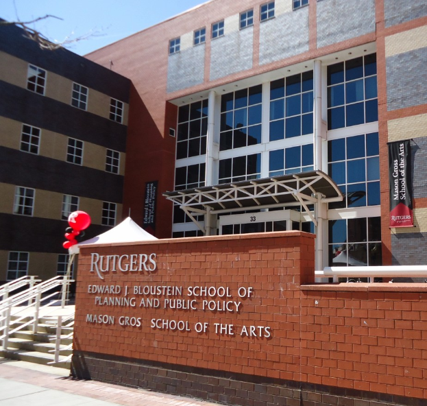 Das Gebäude der Rutgers School of Planning and Public Policy mit Fenstern, Bannern, Treppen mit Geländern, einem Zelt, Luftballons und einem Baum im Vordergrund vor einem klaren blauen Himmel.
