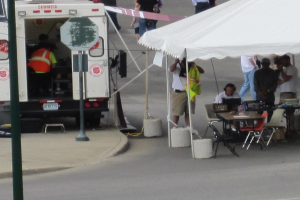 Ein Rettungswagen steht auf der linken Seite einer Straße, während auf der rechten Seite ein Zelt viele Menschen schützt, mit einem Tisch und einem Stuhl in der Nähe.