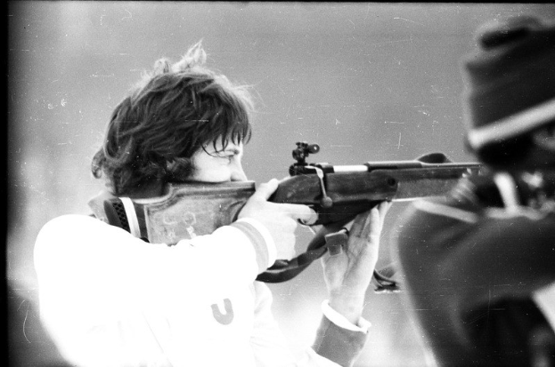 Ein Mann in einem weißen Hemd zielt mit einem Gewehr auf ein Ziel, während rechts neben ihm eine weitere Person steht.