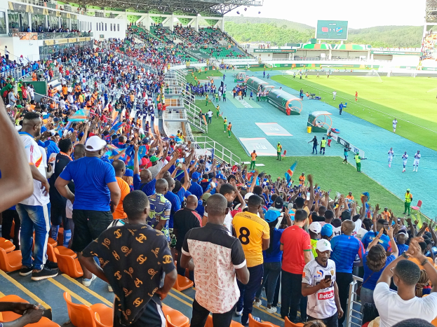Eine große Menschenmenge versammelt sich vor einem eingezäunten Fußballfeld, einige sitzen auf Stühlen, andere stehen, unter einem klaren blauen Himmel mit Hügeln im Hintergrund und warten auf ein Spiel während der Afrika-Cup 2019.