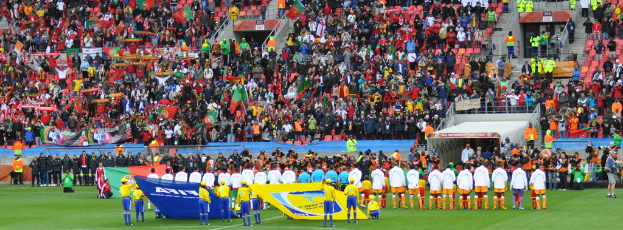 Eine Gruppe von Menschen, die auf dem Boden stehen, einige halten Schilder und Fahnen, mit einem Publikum im Hintergrund, das sitzt und steht, und ein paar Kameraleute auf der rechten Seite.