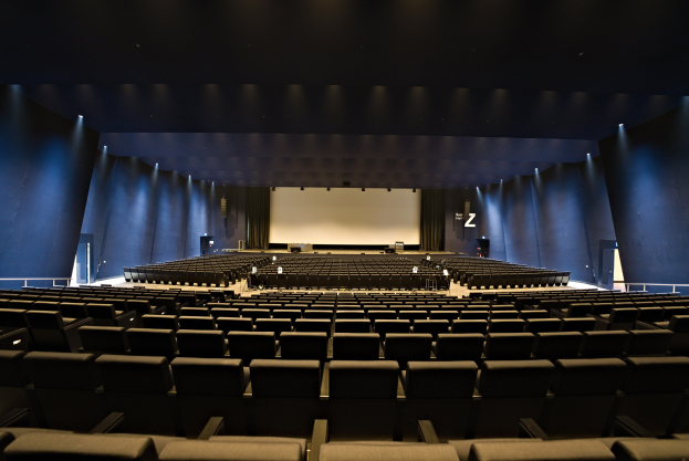 Großer Zuschauerraum mit Sitzreihen, Projektionsbildschirm und Bühnenobjekte, beleuchtet von Wandlampen.