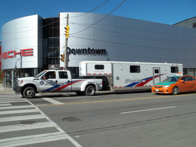 Polizeilkw vor einem Gebäude mit Text geparkt, mit einer Ampel und Kabeln im Vordergrund, Bäume und einen klaren blauen Himmel im Hintergrund.