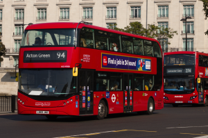Zwei rote Doppeldeckerbusse fahren auf einer von hohen Gebäuden gesäumten Straße, mit Menschen im Inneren der Busse und einem Geländer auf der linken Seite der Straße, mit Bäumen und einem Laternenpfahl im Hintergrund.
