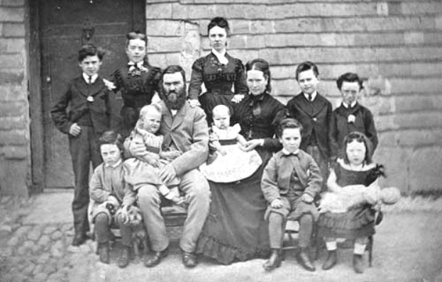 Schwarzes und weißes Foto einer Familie, die vor einem Haus posiert, wobei einige auf einer Bank sitzen und andere stehen; eine Wand und eine Tür sind im Hintergrund zu sehen.