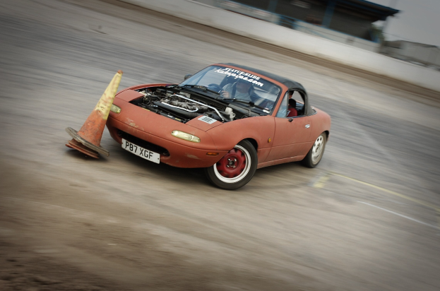 Eine Person fährt ein rotes Auto auf einer Rennstrecke mit einem Verkehrskegel vorneweg, Hintergrund unscharf, was auf hohe Geschwindigkeit hindeutet.