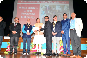 Eine Gruppe von Männern steht auf einer Bühne, zwei halten ein Zertifikat, mit einem Bildschirm im Hintergrund, der 'Indian Institute of Food Processing Technology Awards 2019' anzeigt, und Blumenschmuck auf der Bühne.