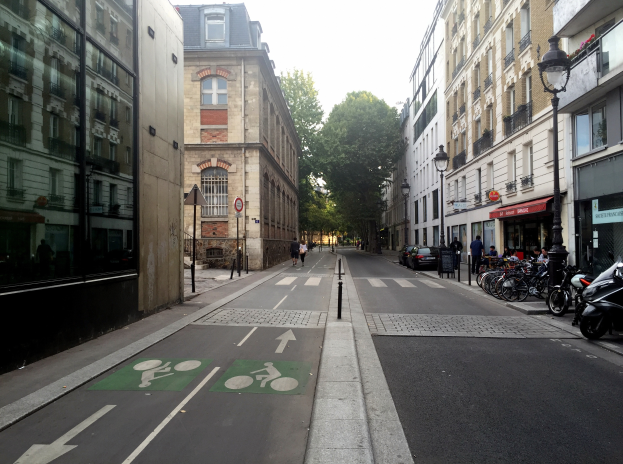 Eine Straße in Paris mit einem Fahrradweg, Fahrzeugen auf der Straße, Fußgängern auf dem Gehweg, Gebäuden mit Fenstern auf beiden Seiten, Bäumen und einem klaren blauen Himmel im Hintergrund.