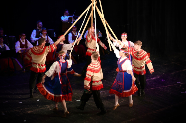 Eine Gruppe von Menschen in farbenfrohen Kostümen, die um einen Maibaum tanzen, einige halten hölzerne Stäbe, mit Musikern im Hintergrund.