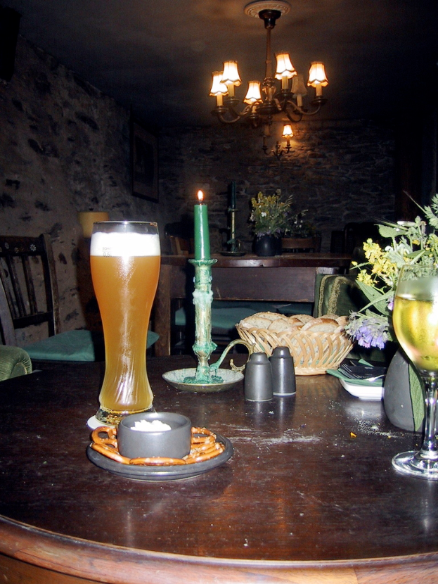 Ein Holztisch in einem Restaurant mit einer Schüssel, einem Glas, einem Korb, einem Weinglas und einer brennenden Kerze, mit anderen Tischen, einer Wand und einem Kronleuchter im Hintergrund.