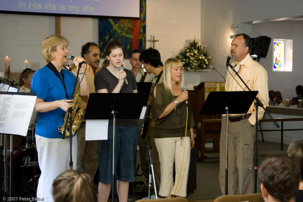 Eine Gruppe von Menschen in einem Raum mit einer Wandbanner, Kerzen, Papier, Mikrofonen, einem Foto Rahmen und Blumen.
