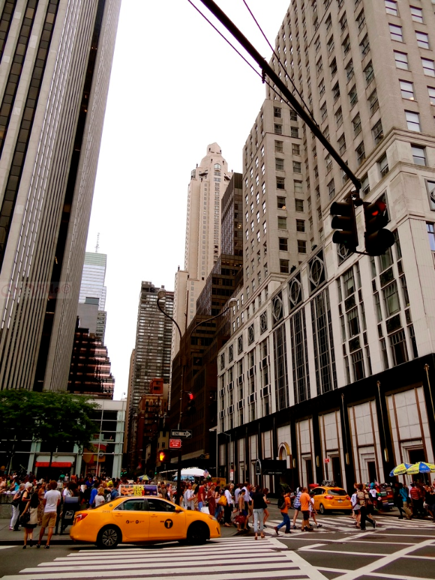 Eine Stadtansicht mit Gebäuden, Ampeln, Fenstern, einigen Passanten, Autos und Himmel.