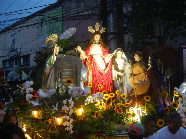 Eine Gruppe von Menschen um einen Umzugswagen herumstehen, der mit Jesus-Statuen, Blumen, Blättern und Lichtern geschmückt ist, mit Gebäuden, Bäumen, Pfählen, Drähten und einem klaren blauen Himmel im Hintergrund.