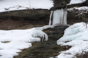 Ein kleiner Wasserfall ergießt sich über einen schneebedeckten, felsigen Hang in einem bewaldeten Gebiet, mit Eiszapfen an den Felsen und schneebedeckten Bäumen, die die Szene umgeben.