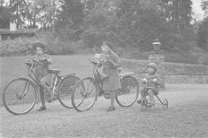 Drei Kinder fahren Fahrräder in einem Park, tragen Mäntel und Mützen und haben eines auf einem Dreirad, vor einem Hintergrund aus Bäumen, Pflanzen, einem Haus und einem Laternenpfahl, in Schwarz-Weiß.