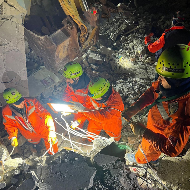 Gruppe von Rettungskräften in orangefarbenen Uniformen und grünen Helmen, die Trümmerschutt von einem eingestürzten Gebäude mit einem Kran im Hintergrund beseitigen.