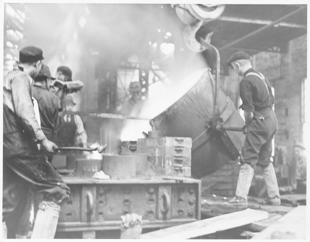 Eine Gruppe von Männern in einer schwarz-weißen Fabrik, die an einem großen Metallobjekt arbeiten, wobei einige Werkzeuge halten und verschiedene Geräte im Hintergrund zu sehen sind.