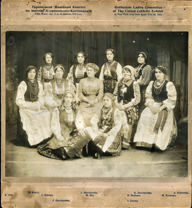 Eine Gruppe von Frauen in traditioneller russischer Kleidung posiert für ein Foto, wobei einige auf dem Boden sitzen und andere stehen, vor einem Vorhanghintergrund, mit Text oben und unten auf dem Bild.