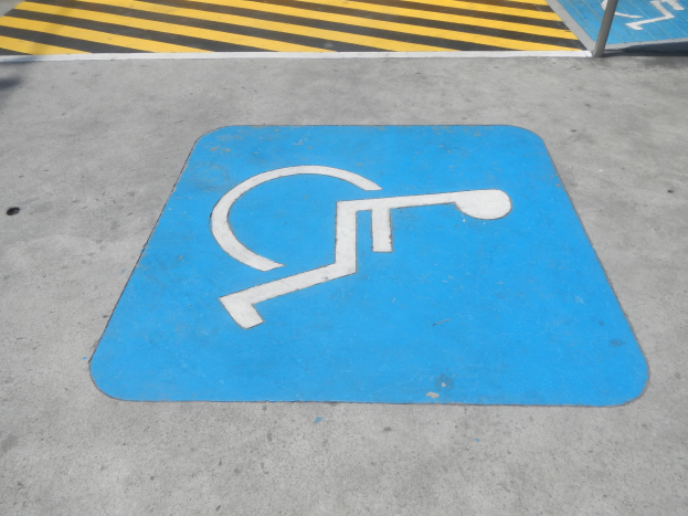 Blaues Behindertenparkplatz-Symbol auf dem Boden mit einer Stange auf der rechten Seite und gelb-schwarzem gestreiftem Boden im Hintergrund.