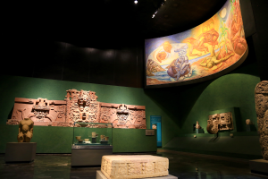 Ein Raum im Mexiko-Stadt-Museum der Anthropologie, gefüllt mit verschiedenen Artefakten, darunter Skulpturen, Gegenstände auf Tischen, eine Texttafel, eine Wandmalerei und Deckenleuchten.